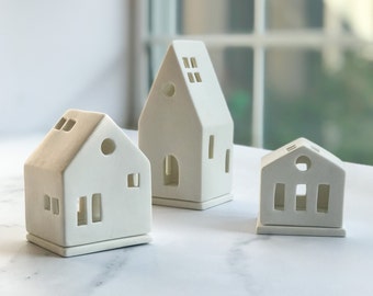 May include: Three off-white ceramic house-shaped candle holders. Each has cut-out windows and doors, with varying rooflines. The smallest has a flat roof, while the others have pitched roofs. The candle holders are arranged on a white surface.