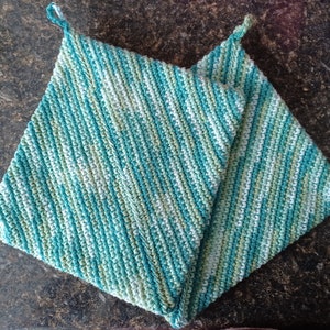 Puede incluir: Dos agarraderas de olla de ganchillo en un esquema de color verde azulado y verde. Las agarraderas de olla están dobladas por la mitad y tienen un patrón diagonal.