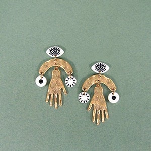 May include: Gold tone earrings with a hand and eye design. The earrings feature a small eye charm, a hand charm, and a larger eye charm.