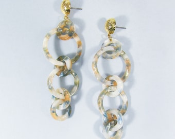 May include: A pair of long, dangling earrings. Each earring features a series of interlocking, marbled resin rings in shades of cream, orange, and blue. The earring posts and top accents are gold-colored.