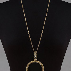 May include: A long gold-tone necklace with a large circular pendant. The pendant is a hollow circle with a decorative beaded element above it. The necklace is displayed on a black mannequin.