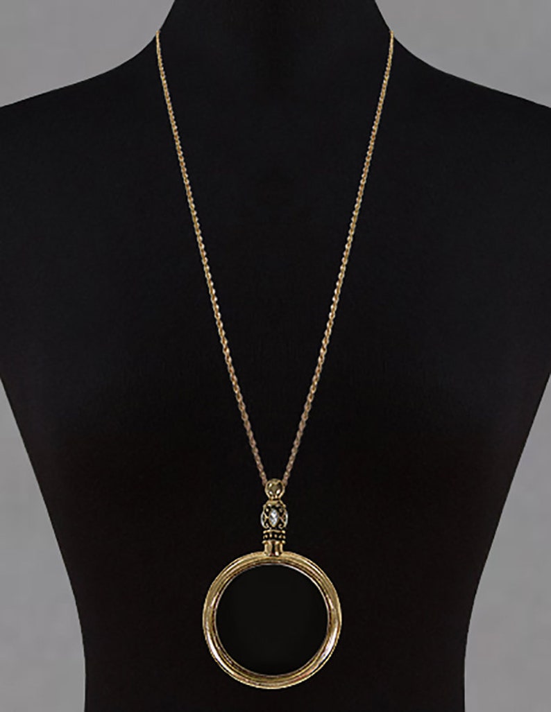 May include: A long gold-tone necklace with a large circular pendant. The pendant is a hollow circle with a decorative beaded element above it. The necklace is displayed on a black mannequin.