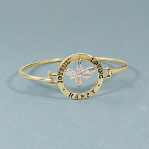 Op de afbeelding: Gouden armband met een bedel van een zilveren bij in het midden van een cirkel met de woorden "Joyful Loving Happy" rond de rand.