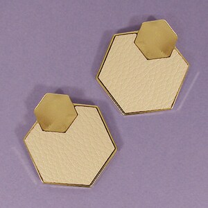 May include: A pair of beige leather earrings with gold geometric accents. The earrings are shaped like a hexagon with a smaller gold hexagon on top.