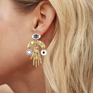 May include: Gold tone earrings with a dangling hand charm, a crescent moon, and an evil eye charm. The charms are white with black details.