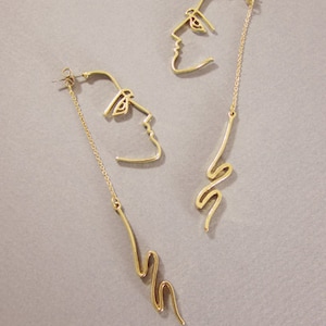 May include: A pair of gold-toned earrings featuring a line drawing of a face on one earring and a wavy line on the other.
