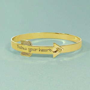May include: Gold bangle bracelet with an arrow charm that says "Follow your heart".
