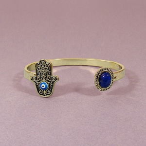 May include: Gold bracelet with a hamsa hand charm featuring a blue evil eye and a blue oval stone.
