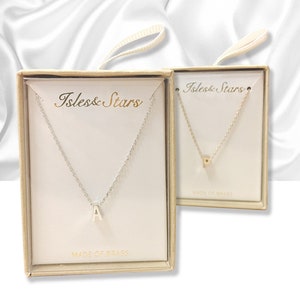 Puede incluir: Dos pequeñas cajas con bordes dorados, cada una contiene un collar de cadena plateada con un pequeño colgante de letra. Las cajas están etiquetadas con "Isles & Stars" y "Made of Brass".