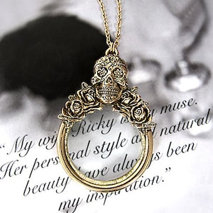 Puede incluir: Un collar dorado con un colgante de lupa. El colgante presenta un diseño detallado de calavera de azúcar con acentos florales. El collar está sobre un fondo blanco con texto que dice "My wife Ricky is my muse. Her personal style and natural beauty have always been my inspiration."
