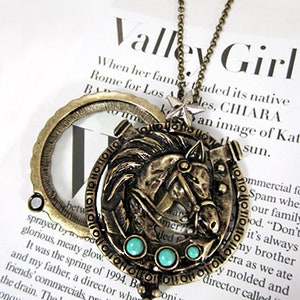 May include: A gold-toned necklace with a magnifying glass pendant featuring a detailed horse design with turquoise accents. The necklace is on a white surface with text that reads "Valley Girl".