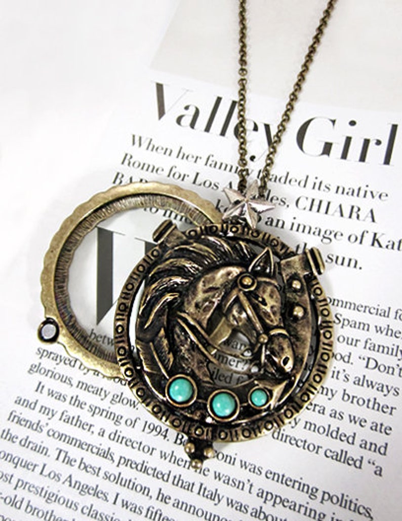 May include: A gold-toned necklace with a magnifying glass pendant featuring a detailed horse design with turquoise accents. The necklace is on a white surface with text that reads "Valley Girl".