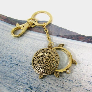 May include: Gold-colored metal keychain featuring a detailed turtle pendant with an openwork design. The keychain includes a lobster clasp and a small chain. The turtle pendant has a clear glass area. The overall design is ornate and decorative.