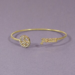 Puede incluir: Un brazalete de oro con un dije de signo de paz y la palabra "paz" inscrita en la banda.