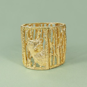 Puede incluir: Un brazalete de metal dorado con un diseño detallado de un gato en una jaula. El brazalete está hecho de una serie de barras de metal conectadas que forman una estructura de jaula. El gato está representado de forma realista, con una cara y un pelaje detallados.