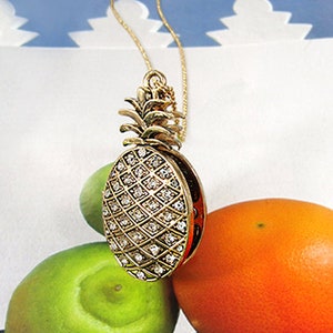 May include: A gold pineapple pendant necklace with clear rhinestones. The necklace is on a white surface with green and orange fruit.