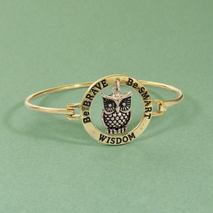 May include: Gold-toned bangle bracelet with a circular charm featuring an owl pendant. The charm is inscribed with "Be Brave, Be Smart, Wisdom." The owl pendant is silver-colored with detailed feathering. The bracelet is set against a green background.