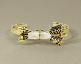 May include: Gold cuff bracelet with two arrowheads on either side of a clear quartz crystal.