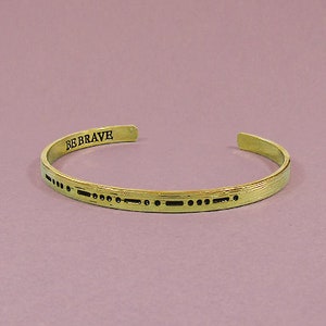 May include: A gold-toned bangle bracelet with the words "BE BRAVE" engraved on the outside. The bracelet is decorated with a pattern of small dots and dashes.