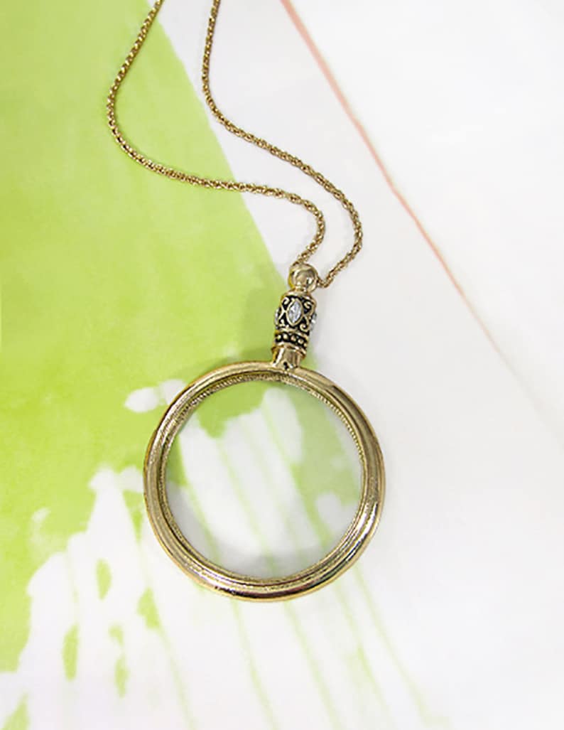 May include: A gold-colored magnifying glass pendant necklace. The pendant features a clear glass lens within a circular frame. The chain and frame have an antique finish. The top of the pendant has a decorative embellishment with a small clear stone.