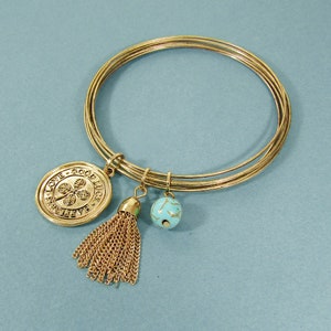 Peut inclure: Bracelet jonc en or avec plusieurs breloques, dont un charme rond avec les mots "Love, Good Luck, Faith" et un trèfle à quatre feuilles, un charme en forme de pompon doré et une perle bleu turquoise.