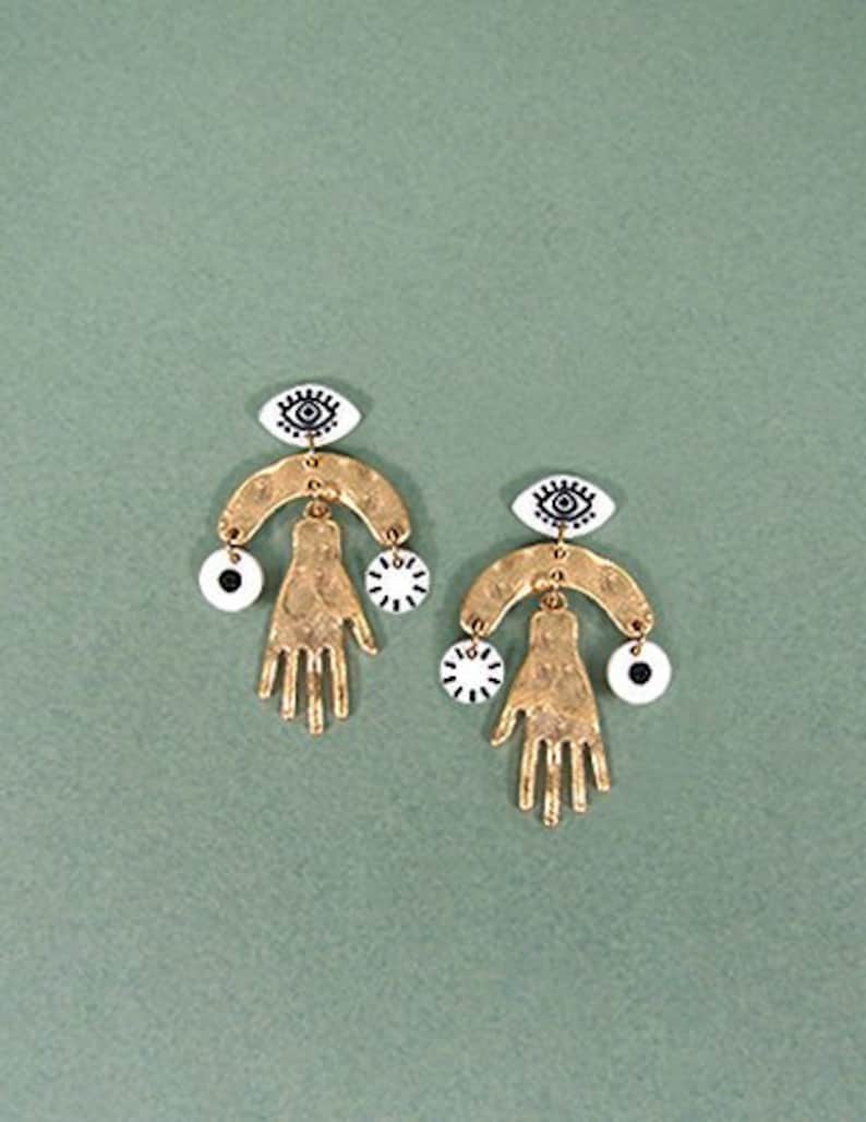 May include: Gold tone earrings with a hand and eye design. The earrings feature a small eye charm, a hand charm, and a larger eye charm.
