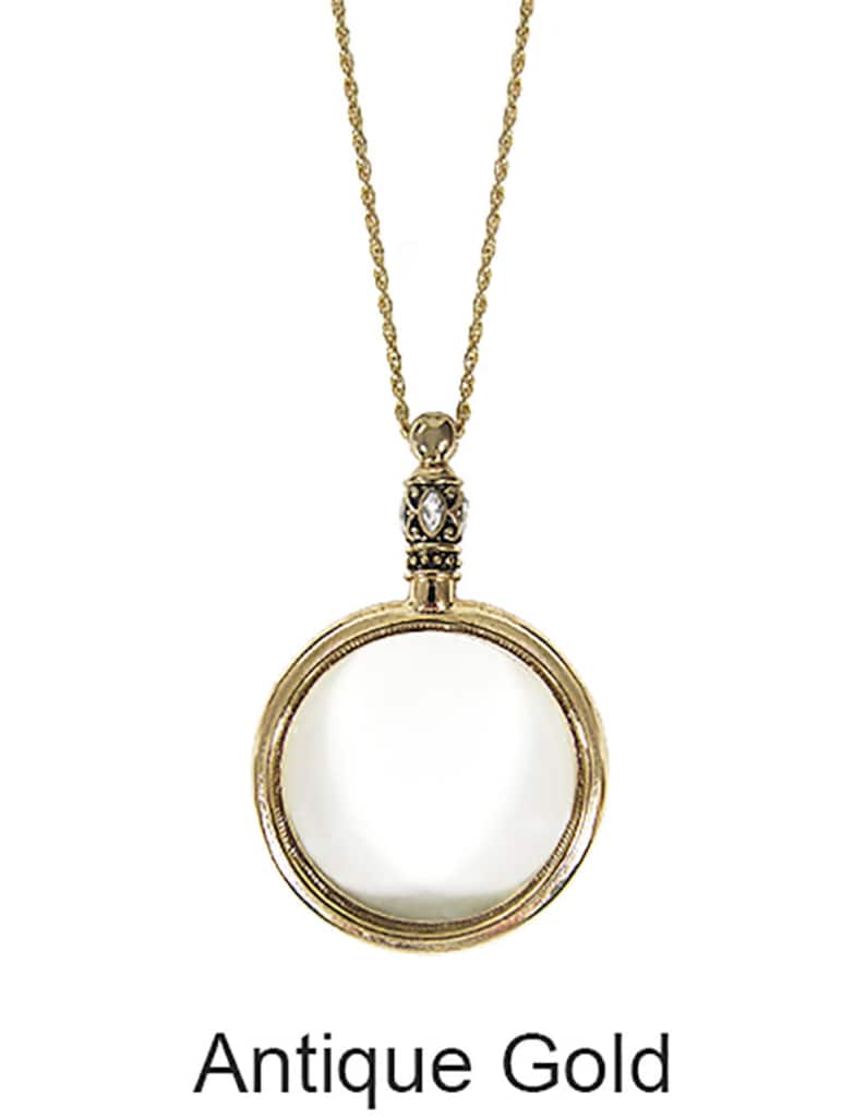 May include: Antique gold-colored magnifying glass pendant necklace. The pendant features a round magnifying glass with a decorative bezel and a small, ornate top with a clear stone. The chain is a delicate gold color. The text "Antique Gold" is below the pendant.