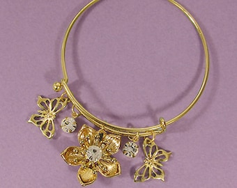May include: Gold-tone bangle bracelet with flower and butterfly charms. The bracelet features a large flower charm with a central clear crystal, two butterfly charms, and two smaller crystal charms. The bracelet is set against a purple background.