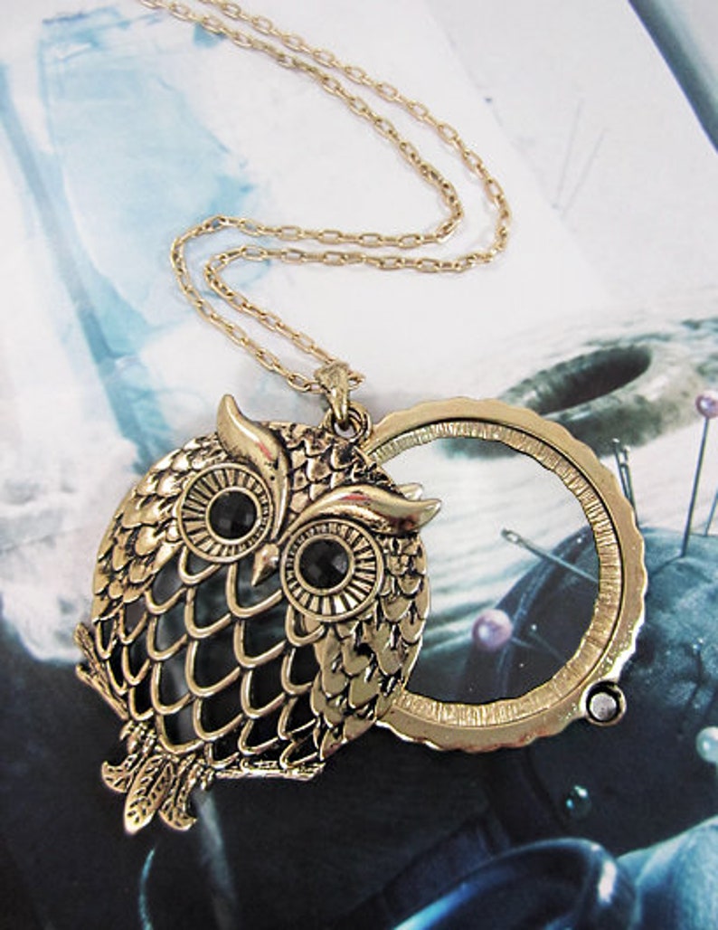 Peut inclure: Un collier avec un pendentif en forme de hibou dor&eacute; avec des yeux en pierres pr&eacute;cieuses noires et un charme de loupe. Le hibou est d&eacute;taill&eacute; avec des motifs de plumes complexes.