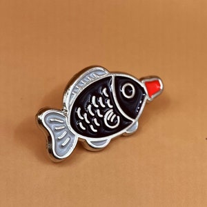 May include: A black and white enamel pin shaped like a fish with a red mouth. The fish has a silver outline.