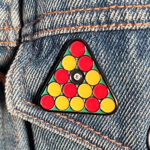 Può includere: Spilla triangolare in smalto con un gruppo di palle da biliardo, tra cui la palla nera numero 8. La spilla ha un bordo verde ed è applicata su una giacca di jeans.