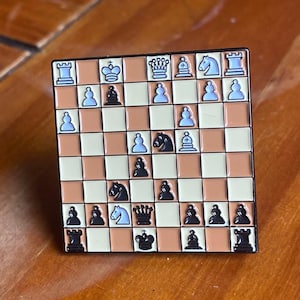 Puede incluir: Pin cuadrado de esmalte con diseño de tablero de ajedrez. El tablero es de color beige y blanco con piezas de ajedrez negras. El pin tiene un borde negro y está sobre una superficie de madera.