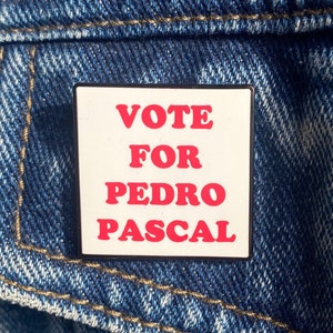 Op de afbeelding: Een vierkante speld met de tekst "VOTE FOR PEDRO PASCAL" in rode letters op een witte achtergrond. De speld is bevestigd aan een blauw spijkerjasje.