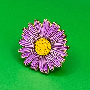May include: A purple and yellow enamel pin featuring a stylized daisy flower.