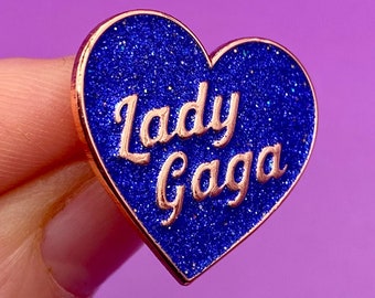 May include: A blue glitter heart-shaped pin with a rose gold outline and the text "Lady Gaga" in rose gold.