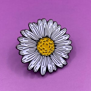 May include: A white daisy enamel pin with a yellow centre. The pin has a black outline and is on a purple background.
