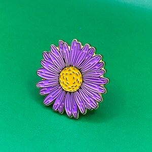 May include: A gold-tone enamel pin featuring a purple flower with a yellow center. The flower has a detailed design and is perfect for adding a touch of whimsy to your clothing or bag.