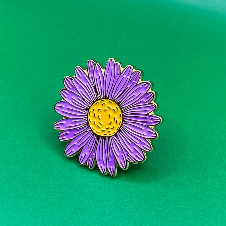 May include: A gold-tone enamel pin featuring a purple flower with a yellow center. The flower has a detailed design and is perfect for adding a touch of whimsy to your clothing or bag.