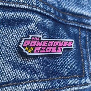May include: A rectangular enamel pin with the text "THE POWERPUFF GIRLS" in pink, outlined in black and white. The design includes two yellow stars and pink hearts. The pin is displayed on a blue denim background.