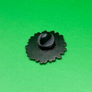 May include: The back of a black enamel pin with a detailed design. The pin has a black metal clasp.