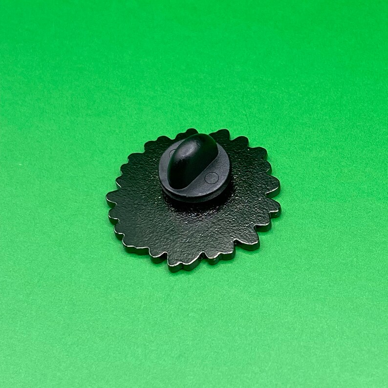 May include: The back of a black enamel pin with a detailed design. The pin has a black metal clasp.