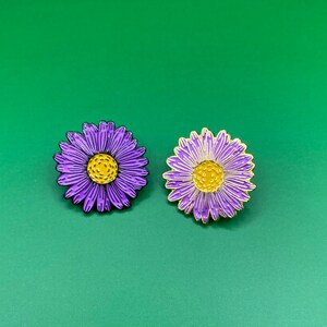 May include: Two purple and yellow enamel flower pins. The flowers have a yellow center and purple petals.
