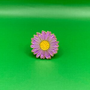 May include: A purple and yellow enamel pin featuring a daisy flower design.