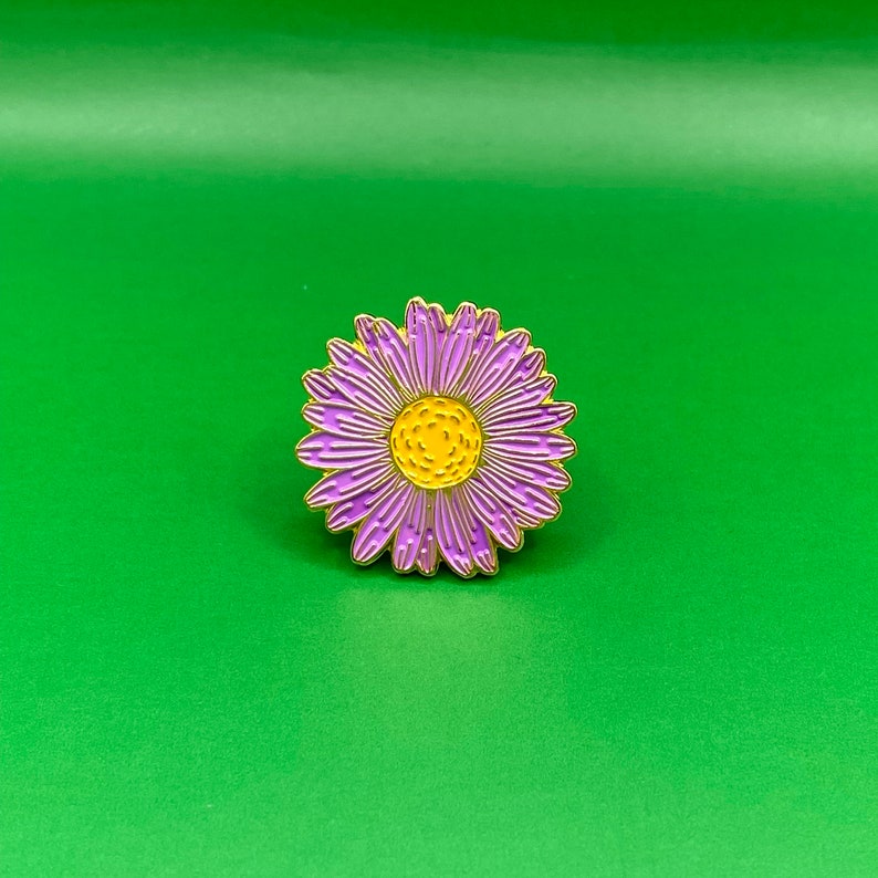 May include: A purple and yellow enamel pin featuring a daisy flower design.