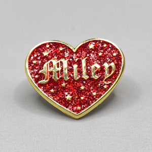 May include: Red glitter heart-shaped pin with gold trim and the word "MILEY" in gold lettering.