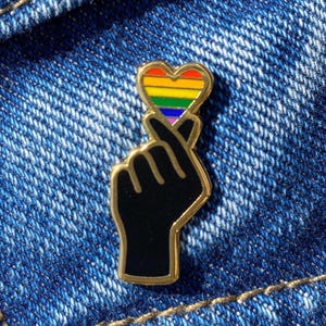 May include: A gold-colored pin featuring a black hand making a heart shape with its fingers. The heart is filled with a rainbow design. The pin is set against a blue denim background.