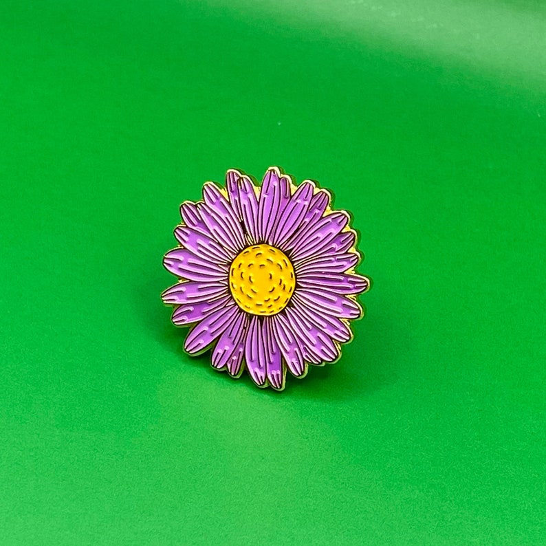 May include: A purple and yellow enamel pin featuring a stylized daisy flower.