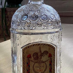 May include: A clear glass bottle with a textured surface and a label that reads "eau de cologne du COQ GUERLAIN 68, Champs-Elysees, PARIS".