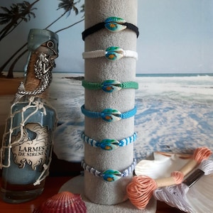 Peut inclure: Six bracelets en cordon tressé empilés sur un présentoir gris, chacun doté d'un coquillage cauri central peint avec un motif de tortue de mer verte et bleue. Les bracelets sont noirs, blancs, verts, bleus, bleu clair et à motif chevron.