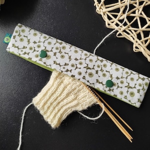 Könnte beinhalten: Ein Stricknadel-Etui mit weißem und grünem Blumenmuster und zwei grünen Knöpfen. Ein kleines, cremefarbenes Strickmuster und Stricknadeln aus Holz sind ebenfalls zu sehen. Das Etui ist geöffnet und zeigt eine grüne Innenseite.